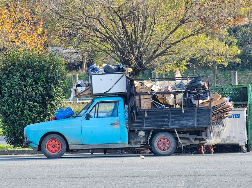 A small, vintage blue flatbed truck parked on the side of a street, fully loaded with various types of rubbish and waste materials. The vehicle's body shows signs of wear with faded paint and minor rust, and the red-painted wheels contrast with the blue body. The truck bed is filled with a mixture of cardboard boxes, black plastic bags, wooden pallets, and other debris, all piled high and secured with ropes or cords. In the background, there is a row of trees with autumn-colored foliage, and green fencing alongside the roadside. The scene appears to be an independent rubbish collection or on-site clearance operation, with the waste awaiting disposal. The natural lighting highlights the textures of the materials and the surroundings, emphasizing the unorganized nature of the load, consistent with private waste handling services highlighting rubbish removal tasks near residential or public areas.