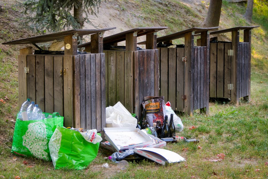 A small, vintage blue flatbed truck parked on the side of a street, fully loaded with various types of rubbish and waste materials. The vehicle's body shows signs of wear with faded paint and minor rust, and the red-painted wheels contrast with the blue body. The truck bed is filled with a mixture of cardboard boxes, black plastic bags, wooden pallets, and other debris, all piled high and secured with ropes or cords. In the background, there is a row of trees with autumn-colored foliage, and green fencing alongside the roadside. The scene appears to be an independent rubbish collection or on-site clearance operation, with the waste awaiting disposal. The natural lighting highlights the textures of the materials and the surroundings, emphasizing the unorganized nature of the load, consistent with private waste handling services highlighting rubbish removal tasks near residential or public areas.
