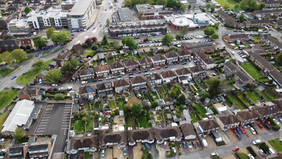 An aerial view of a residential area showing closely spaced terraced houses with small, well-maintained gardens at the rear, separated by narrow pathways and fences. The houses have pitched roofs with dark tiles, some featuring solar panels, and are painted in neutral tones. Cars are parked along the narrow streets and driveways in front of the houses, with a variety of vehicles visible. Surrounding the residential zone are patches of greenery, including mature trees and lawns, as well as a larger commercial or institutional building with a parking lot at the edge of the scene. The environment appears orderly and typical of suburban housing, with natural daylight enhancing the visibility of details. This aerial perspective provides insight into a typical UK neighborhood, useful for understanding urban layout and planning related to rubbish removal services in residential areas aligned with private waste management options like on-site clearance or independent collection offered by companies such as House Clearance Merton.