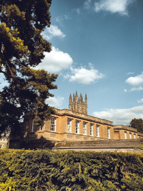 A historic stone building with a prominent clock tower featuring multiple spires, situated in a lush green area with well-maintained bushes and trees in the foreground. The building's exterior walls are made of warm-colored sandstone with detailed architectural elements, and it has large rectangular windows with white frames. A large, dark green tree partially overlaps the left side of the structure, casting shadows on the lower part of the building. The sky above is partly cloudy with scattered white clouds and bright blue patches, creating a natural, daylight ambience. This scene reflects a well-preserved historic site, possibly subject to private or independent waste management practices, aligning with a context of non-local authority rubbish disposal or site clearance, supported by the overall tidy appearance and lack of visible waste or debris.