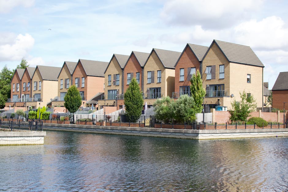 A row of modern, multi-storey residential houses situated along a waterfront, with each building featuring a mix of beige, brown, and reddish brick facades. The houses have pitched grey tiled roofs and small front balconies with glass railings. In the foreground, a calm body of water reflects the buildings and the partly cloudy sky overhead. The properties are separated from the water by a paved promenade bordered by black metal fencing, with some greenery including neatly trimmed trees and bushes planted along the edge. The environment appears clean and orderly, typical of suburban estate developments, and the scene conveys a quiet, residential atmosphere. House Clearance Merton may be involved in managing waste and rubbish collection related to property maintenance or estate clearance services in this area, supporting private disposal or alternate waste handling outside local authority collections.