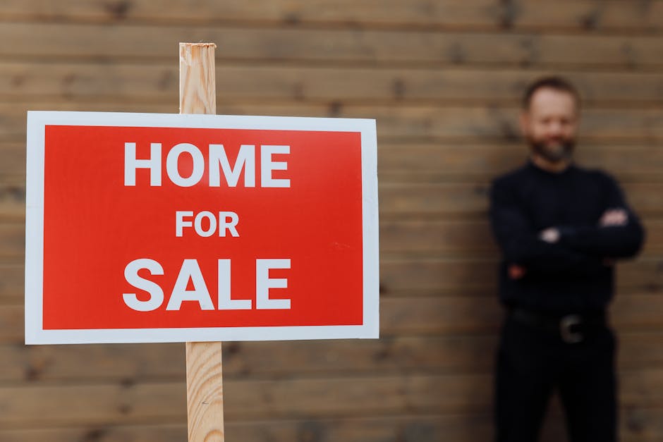 A close-up view of a rectangular 'Home for Sale' sign with a red background and white bold text, mounted on a light wooden stake. The sign is positioned in the foreground of the image, occupying the left side, and is clearly visible. In the background, there is a blurred man with a beard and short hair, dressed in a black shirt and black trousers, standing with arms crossed in front of a wooden slat fence or wall, which has a natural, warm brown tone. The lighting appears natural, illuminating the sign brightly while the background remains softly lit. The scene is set outdoors, possibly on a driveway or garden area adjacent to the property, with an emphasis on the sign indicating a property available for purchase, subtly related to property clearance or estate disposal activities managed by houseservice providers like House Clearance Merton.