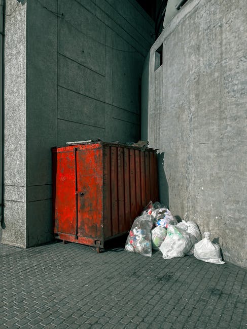 An outdoor urban scene featuring a large red metal refuse container with visible signs of rust and wear, positioned against a dark green wall on the left side and a grey concrete wall on the right. Several black, clear, and white plastic rubbish bags, some partially torn open, are stacked or scattered at the base of the container, spilling onto the paved grey sidewalk made of interlocking bricks. The background includes a narrow alleyway or side street with a small, rectangular window visible high on the grey wall, and the overall environment appears to be a utilitarian area designated for waste collection or disposal, characteristic of private rubbish clearance services provided by companies like House Clearance Merton.
