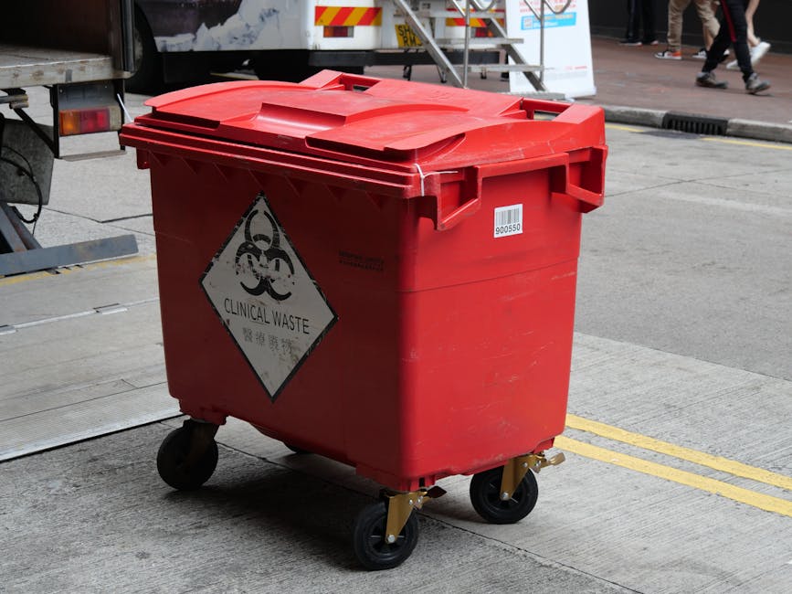A red plastic wheeled bin designated for clinical waste disposal, positioned on a grey concrete pavement next to a vehicle with a partially visible rear. The bin features a black and white hazard symbol with the words 'CLINICAL WASTE' beneath it, indicating its use for medical or hazardous waste. The surface of the bin appears slightly worn with some dirt or scuff marks, and its lid is tightly closed. In the background, part of a truck or container is visible, with a metal staircase and some pedestrians walking on a pathway further behind. The scene suggests an environment where designated waste containers are used for proper disposal, aligning with private rubbish removal or on-site clearance activities that require specialist handling of hazardous waste, as managed by companies like House Clearance Merton.