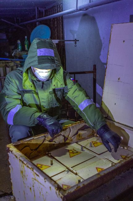 A person wearing a high-visibility green and grey protective suit with reflective stripes, black gloves, and a face mask is crouched down inspecting or handling contents inside an open, large industrial waste container with yellow hazard warning labels on its interior sides. The setting appears to be an indoor storage area or waste facility, with dim lighting and background elements including a shelf with bottles and other objects, as well as a blue plastic item on the wall. The container's exterior is worn and painted in a pale yellow or off-white shade, with visible signs of use. The individual appears to be conducting a detailed examination, possibly as part of a hazardous waste handling or disposal process, in line with private rubbish removal or on-site clearance services often undertaken by companies like House Clearance Merton. The scene emphasizes careful handling and assessment of waste materials, aligning with professional waste management practices for hazardous or specialized rubbish removal.