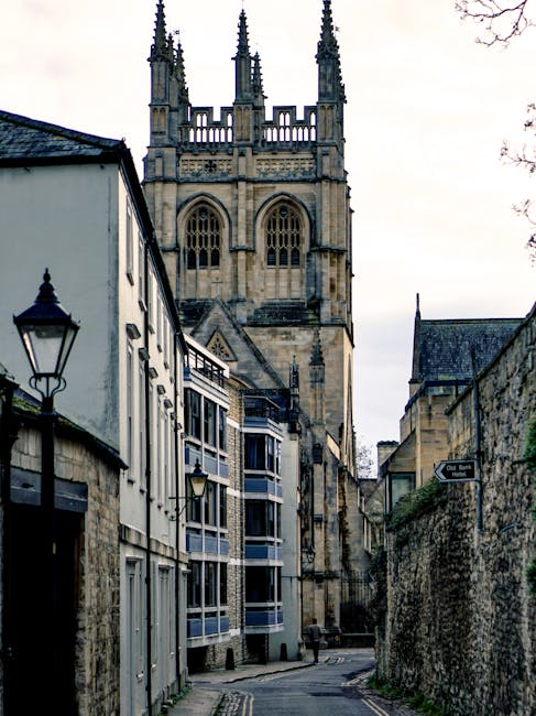 A narrow, winding street in an old town with a mix of modern and historic buildings. On the left, there are white-painted terraced houses with black-framed windows and traditional black metal lamp posts attached to the walls. The buildings have a combination of flat roofs and sloped tiled roofs, with some featuring small balconies. On the right, there is a high, weathered stone wall with a street sign attached, indicating the location of the street. In the background, towering gothic-style church spires with intricate stone detailing reach towards the cloudy sky, dominating the scene and emphasizing the historic architectural character of the area. The street appears quiet and clean, with no visible vehicles or pedestrians, and natural, subdued lighting enhances the textures of the stone and paint, creating a calm, atmospheric setting typical of a historic urban environment where private waste handling and on-site clearance services might be relevant for residents or businesses seeking alternatives to local authority rubbish collection. The scene subtly underscores the importance of proper waste disposal in heritage-rich areas managed by companies like House Clearance Merton, serving local communities with expertise in rubbish removal services.