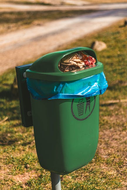 A green public rubbish bin with a semi-transparent oval lid, partially open, revealing assorted waste including paper, plastic, and metal items inside. The bin features a blue plastic liner wrapped around the top rim and has a black recycling symbol printed on its front, indicating waste separation. It is mounted on a metal pole situated outdoors on a grassy area, with a dirt path and blurred greenery in the background under natural daylight. The scene suggests it is in a park or similar communal space, where waste collection is essential, and highlights the importance of proper rubbish disposal consistent with private clearance services provided by House Clearance Merton in the context of local rubbish removal and waste management practices.