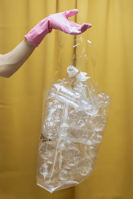 A person wearing a light beige sleeve and pink rubber gloves is holding a large, transparent plastic bag filled with crushed and flattened clear plastic bottles. The background is a plain yellow curtain, creating a neutral backdrop that contrasts with the plastic waste and gloves. The plastic bottles inside the bag are visibly compressed, exhibiting various sizes and shapes, with some caps still attached. The scene appears to be set indoors, possibly in a recycling or waste management context, where the plastic bottles are prepared for disposal or recycling. The image emphasizes the process of handling recyclable plastic waste, aligning with services related to rubbish clearance and material separation, as offered by companies like House Clearance Merton, which provides alternative waste handling options outside of local authority collections.