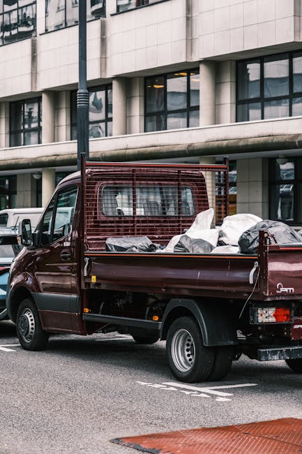 A burgundy flatbed utility vehicle parked on an urban street in front of a modern office building with large glass windows. The truck's cargo area is loaded with various types of rubbish, including black and white plastic bags and miscellaneous debris, ready for collection or disposal. The vehicle features a protective metal grid at the cab's rear for safety, and the wheels are equipped with silver hubcaps. The scene is illuminated by natural daylight, with a street lamp visible above the truck and other parked cars seen in the background. The setting suggests an environment where private waste handling, such as rubbish removal services by companies like House Clearance Merton, is conducted to clear waste from urban properties or commercial premises, emphasizing alternative waste collection methods outside local authority services.