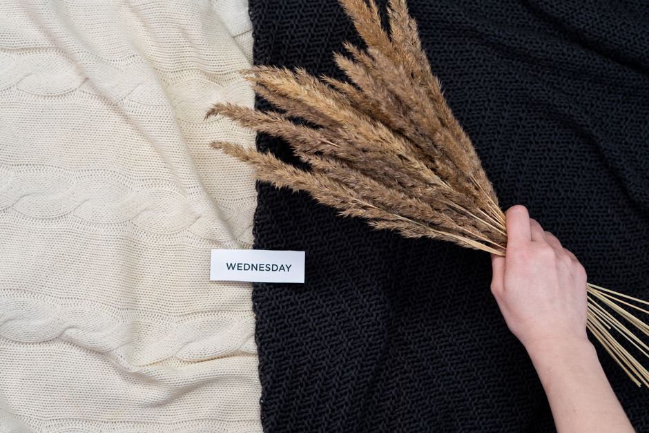 A close-up photograph features a fair-skinned hand holding a small bouquet of dried beige ornamental grass stalks with feathery plumes. The hand is positioned in the lower right corner of the image. The background is divided into two textured fabric surfaces; on the left, a cream-colored knitted material with a pattern of interwoven loops, and on the right, a black knitted fabric with a coarser, more open weave. A small white rectangular label with the word 
