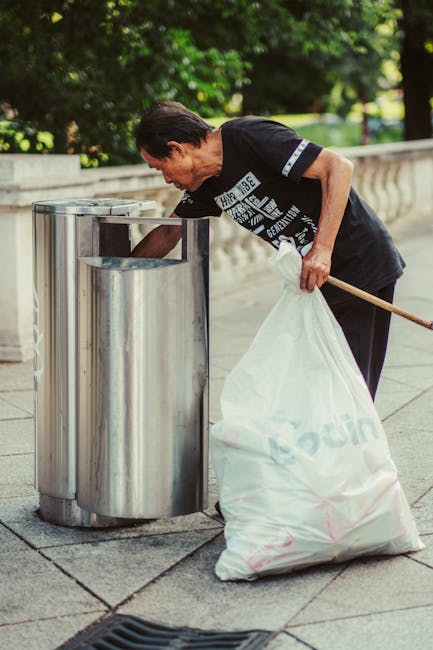 A middle-aged woman with short dark hair is seen disposing of waste into a stainless steel dual-compartment public bin positioned on a paved outdoor walkway. She is wearing a black T-shirt with white text and dark trousers, and is holding a large white plastic bag filled with rubbish in her left hand, while reaching into the bin with her right hand. The bin has a modern, polished finish and is set against a backdrop of lush green trees and landscaped foliage, indicating a park or public garden area. The scene is illuminated by natural daylight, highlighting the textures of the bag, metal, and pavement. The woman's posture and actions suggest an act of waste disposal, which could be part of a community cleanup or individual rubbish removal, supporting the context of private or independent waste handling services, such as those provided by House Clearance Merton.