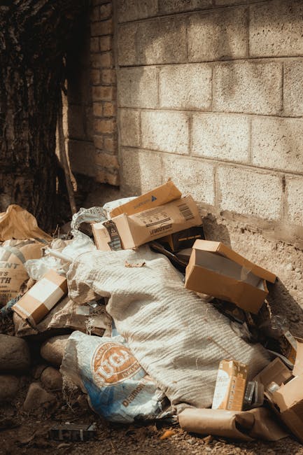 A pile of mixed rubbish consisting of flattened cardboard boxes, plastic bags, and paper wrapping, situated on the ground adjacent to a brick and concrete wall. The boxes are in various shades of brown and grey, with some partially torn or crumpled. A large white sack is visible within the heap, containing additional waste. The surrounding environment appears to be an outdoor area near the corner of a building, with a tree trunk partially visible on the left side of the image. The waste has accumulated on a surface of uneven soil or gravel, with some loose rocks scattered around. The scene suggests an instance of ongoing rubbish accumulation, which could be addressed by on-site or private waste collection services such as those offered by House Clearance Merton, specialising in rubbish removal and clearance activities.