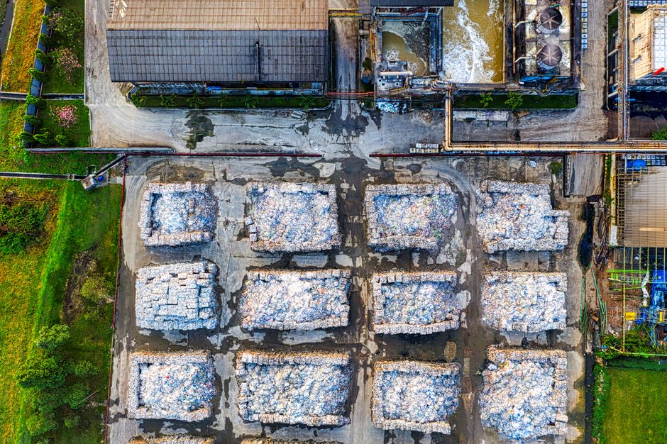 An aerial view of a site containing multiple large piles of white waste materials, likely construction debris or insulation boards, arranged in eight rectangular stacks on a paved surface. The stacks are situated in a semi-organized pattern with visible gaps between them, and the materials have a rough texture with some uneven edges and scattered debris on top. Surrounding the waste piles, there is a mix of dirt and asphalt, with patches of darker wet areas near the top center of the image. To the top left, part of a building with a sloped, dark tiled roof and grey walls is visible, with green plants in a garden area adjacent to a curved driveway or pathway leading to the site. On the right side, there are additional structures, including metallic and wooden elements, and what appears to be some on-site equipment or containers. The overall scene suggests a waste management or disposal operation, possibly related to private rubbish clearance or on-site material handling, consistent with services offered by companies like House Clearance Merton.