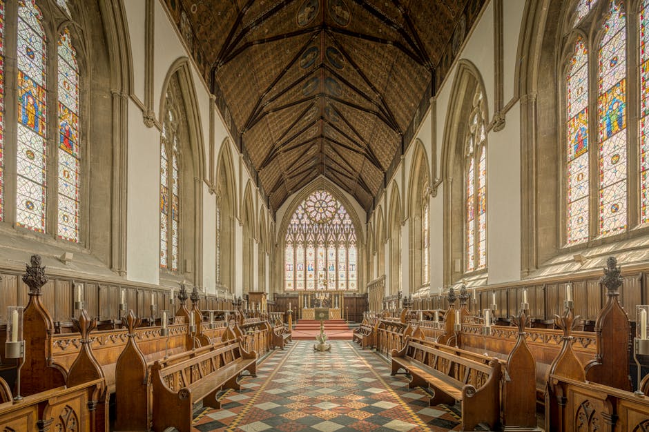 Inside a historic church, the high vaulted wooden ceiling features intricate dark timber beams. Tall, arched stained-glass windows on both sides of the aisle let in soft, colorful light, illuminating the space with a warm glow. The side walls are lined with decorative wooden paneling and ornate candleholders attached to the pews, which are made of polished dark wood with carved details. The central aisle has a patterned tile floor with earthy tones, leading towards the altar at the far end, which is visible through an elaborate, pointed stained-glass window. The altar area is elevated and adorned with religious symbols, with natural light streaming through the large windows behind it. The overall atmosphere suggests a peaceful, historic place of worship with carefully maintained wooden furnishings and stained-glass artistry, subtly evoking themes of preservation and careful space management typical of private or independent arrangements, aligning with the concept of alternative waste handling and discreet clearing services offered by House Clearance Merton.