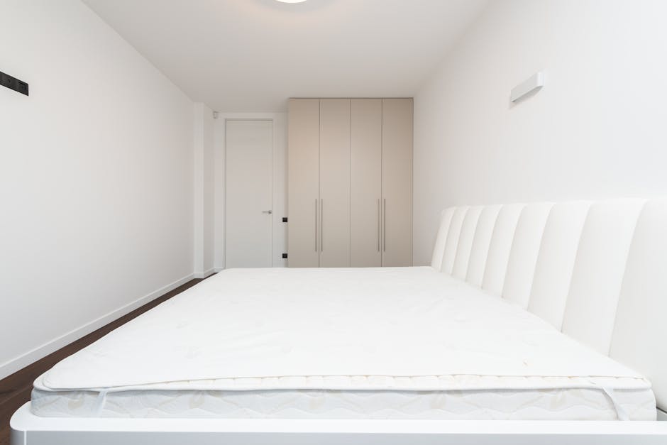 A spacious, well-lit bedroom featuring a large bed with an off-white, tufted mattress and matching upholstered headboard positioned on the right side of the image. On the nightstand beside the bed, there is a modern, cream-colored table lamp with a rounded base. To the left of the bed, a light-colored armchair is placed near a window with sheer white curtains, allowing natural light to fill the room. A small, round glass-top side table is situated in front of the window, and a stylish, slim black desk with a matching chair is positioned against the left wall, beneath a rectangular mirror. The room has neutral-toned walls with crown molding along the ceiling, and the floor is hardwood with a polished, smooth finish. The overall atmosphere is airy and minimalist, with unobstructed views of greenery outside the window, reflecting a quiet, modern interior that might be targeted for private or on-site waste clearance services by House Clearance Merton when preparing a property for rubbish removal including mattress disposal.
